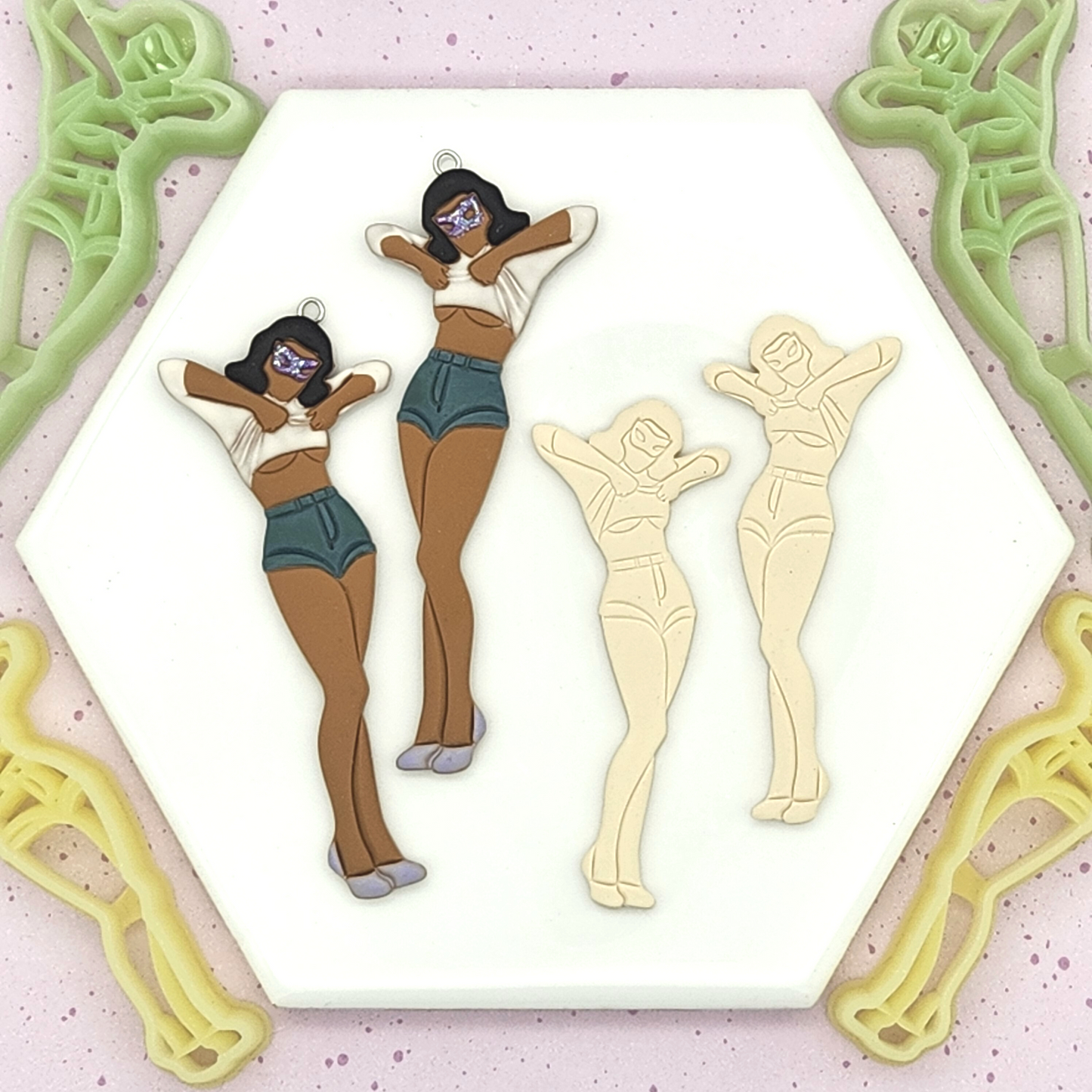 two fully decorated clay pieces of a woman with dark hair, sunglasses, a white top, and high-waisted denim shorts, alongside two plain clay cutouts. Green and yellow cutters in the same pose surround them.