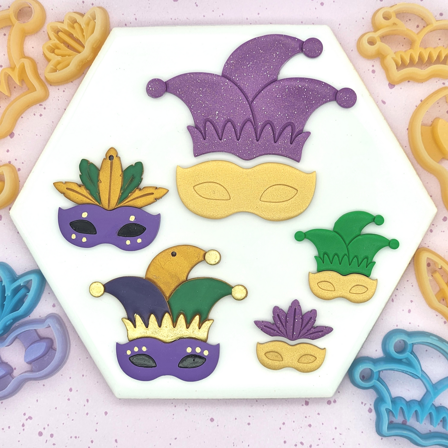 A bold set of Mardi Gras–inspired polymer clay pieces laid out on a white hexagon tile, featuring jester hats and masks in purple, green, and gold, surrounded by matching cutters.