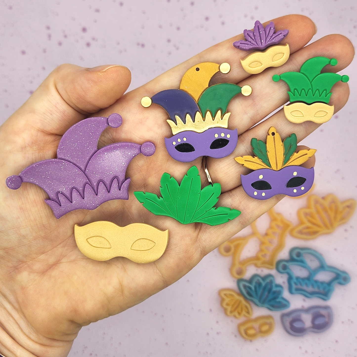 Hand displaying multiple miniature jester hats and masks in vibrant Mardi Gras colors, showing off the scale of each finished polymer clay piece