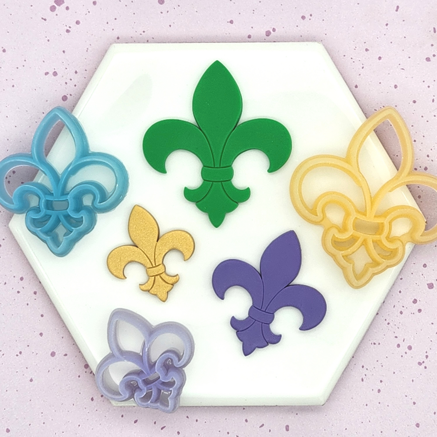 A collection of fleur-de-lis-shaped polymer clay cutters and clay pieces in various colors, displayed on a hexagonal white tile. The cutters come in blue, yellow, and purple, while the clay pieces are green, gold, and purple