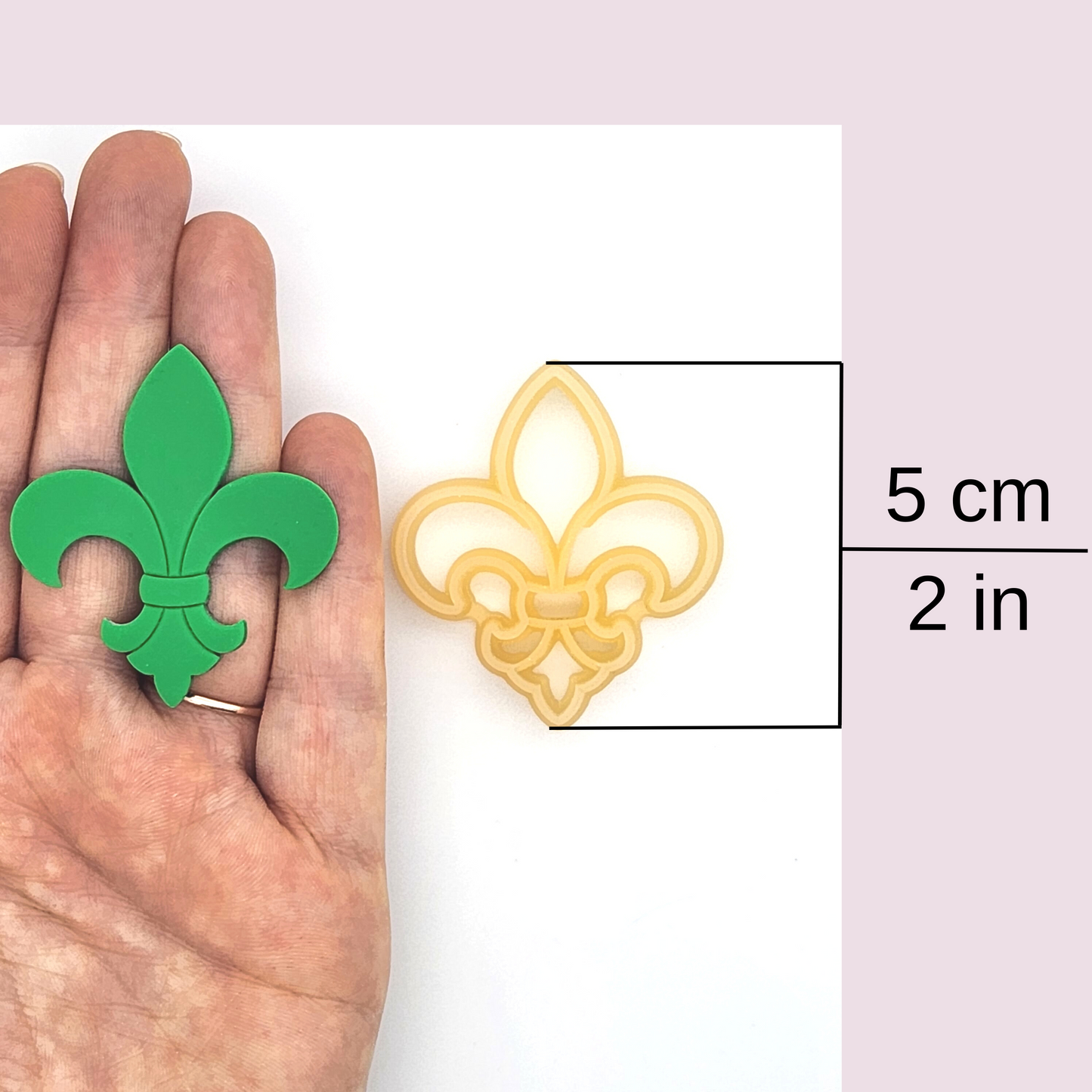 a hand holding a green clay fleur-de-lis and a yellow cutter with a size label indicating 5 cm (2 in)
