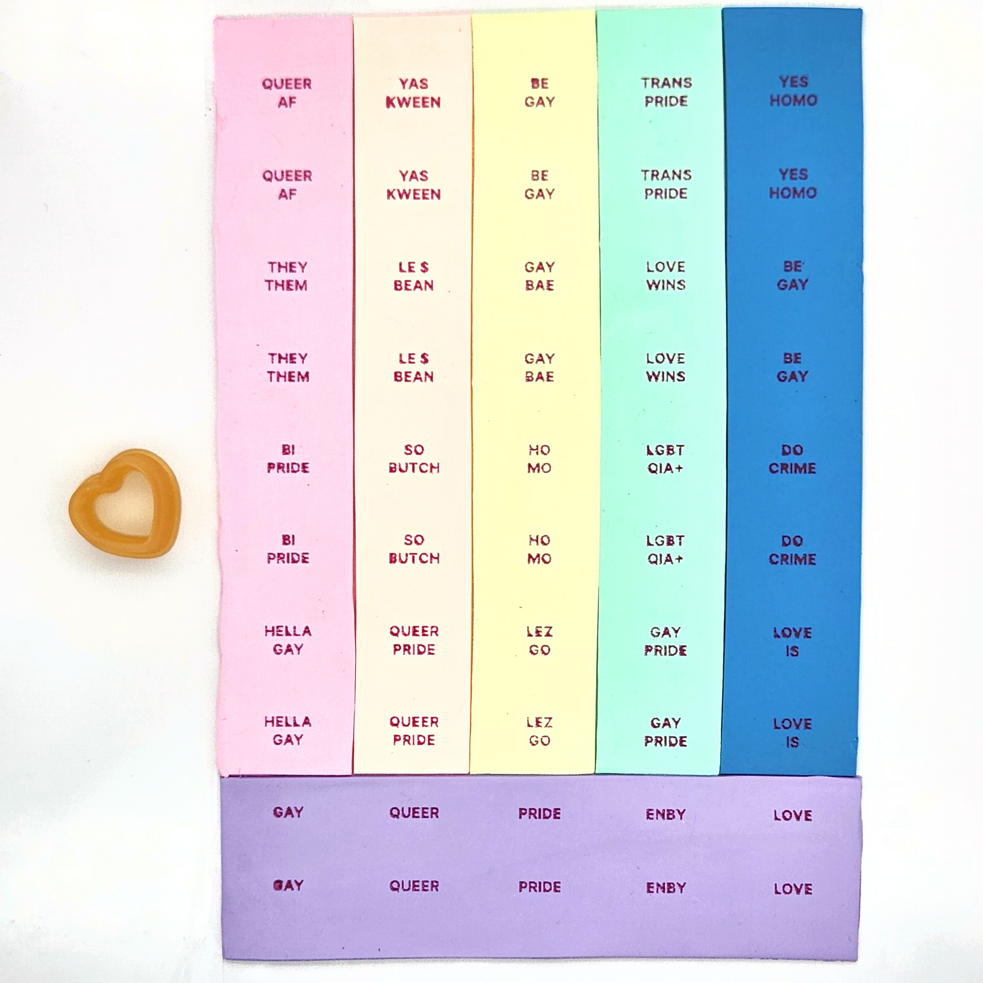 queer themed words painted on clay: "HELLA GAY", "QUEER AF", "THEY/THEM", "BI PRIDE", on pink polymer clay. The words "QUEER PRIDE", "SO BUTCH", "YAS KWEEN", "LE$ BEAN" on orange polymer clay. The words "GAY BAE", "BE GAY", "HO MO", "LEZ GO" on yellow pol