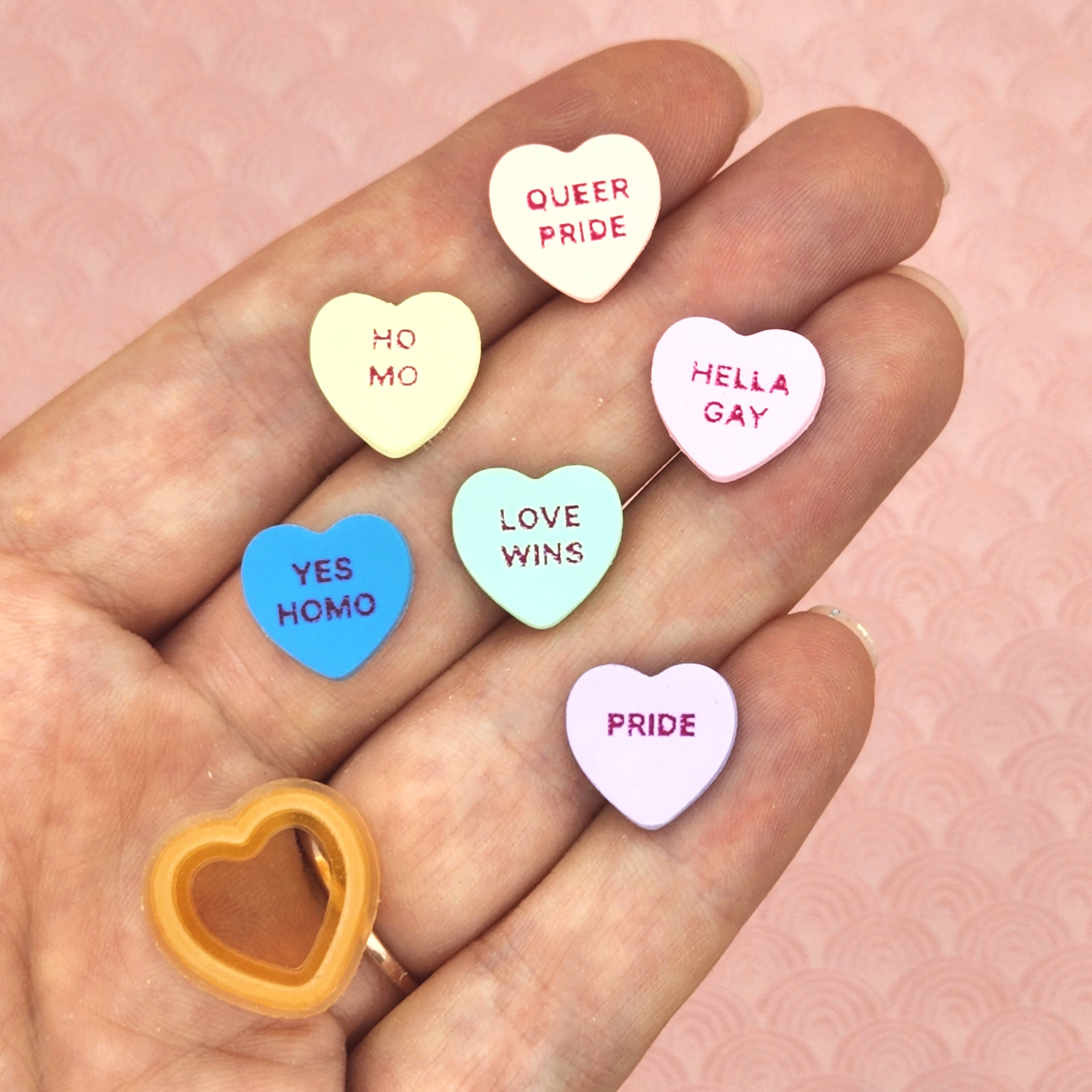 polymer clay conversation hearts with the words "QUEER PRIDE", "HO MO", "HELLA GAY", "LOVE WINS", "YES HOMO", "LOVE WINS", "PRIDE"